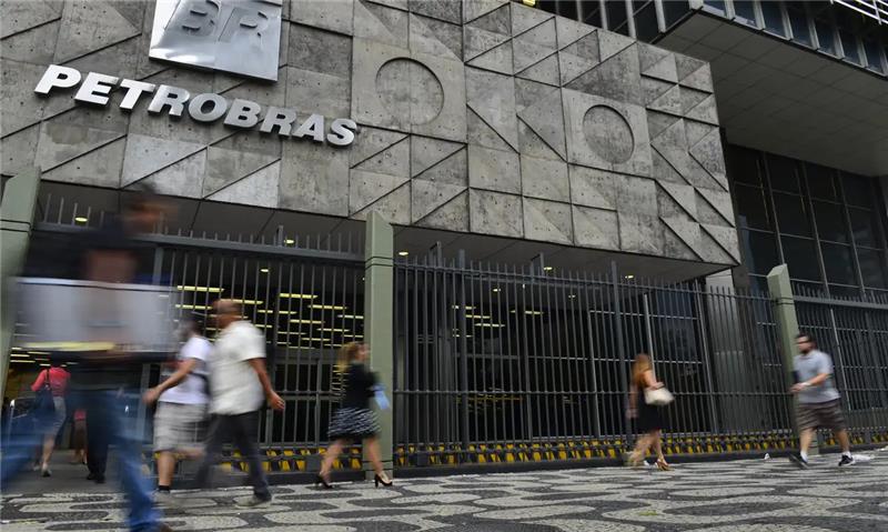 Fachada do edifício-sede da Petrobras, no centro do Rio de Janeiro. A imagem mostra a entrada principal, com o logotipo da empresa em letras metálicas na parte superior e um painel de concreto geométrico com formas triangulares e circulares. Em primeiro plano, várias pessoas caminham em frente ao prédio, algumas desfocadas pelo movimento. O piso é de pedras portuguesas em preto e branco, formando desenhos ondulados típicos das calçadas da cidade.