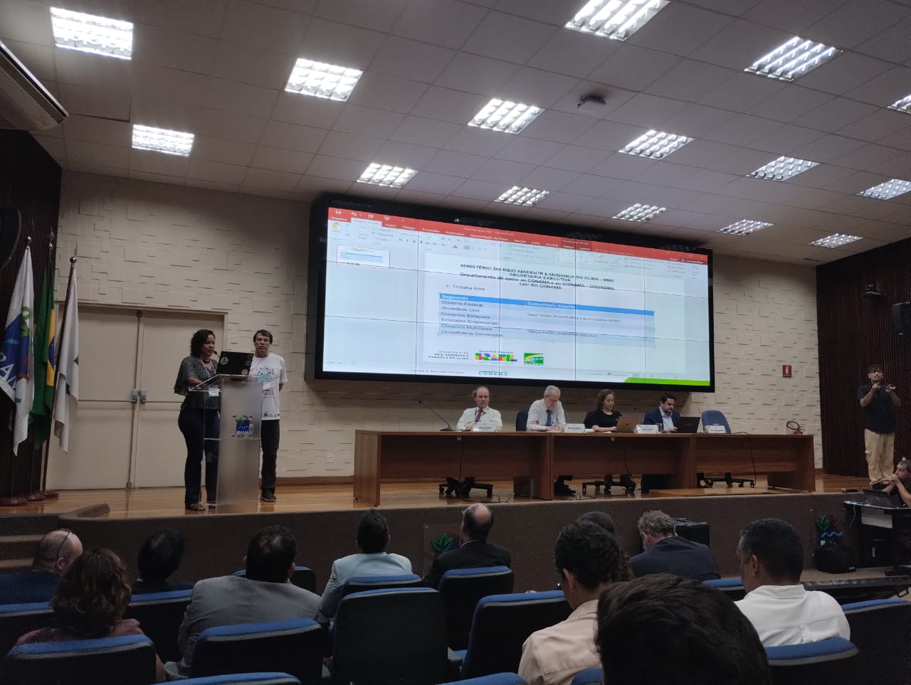 2 Pessoas em um palco com uma tela ao fundo contendo uma apresentação em ppt.
