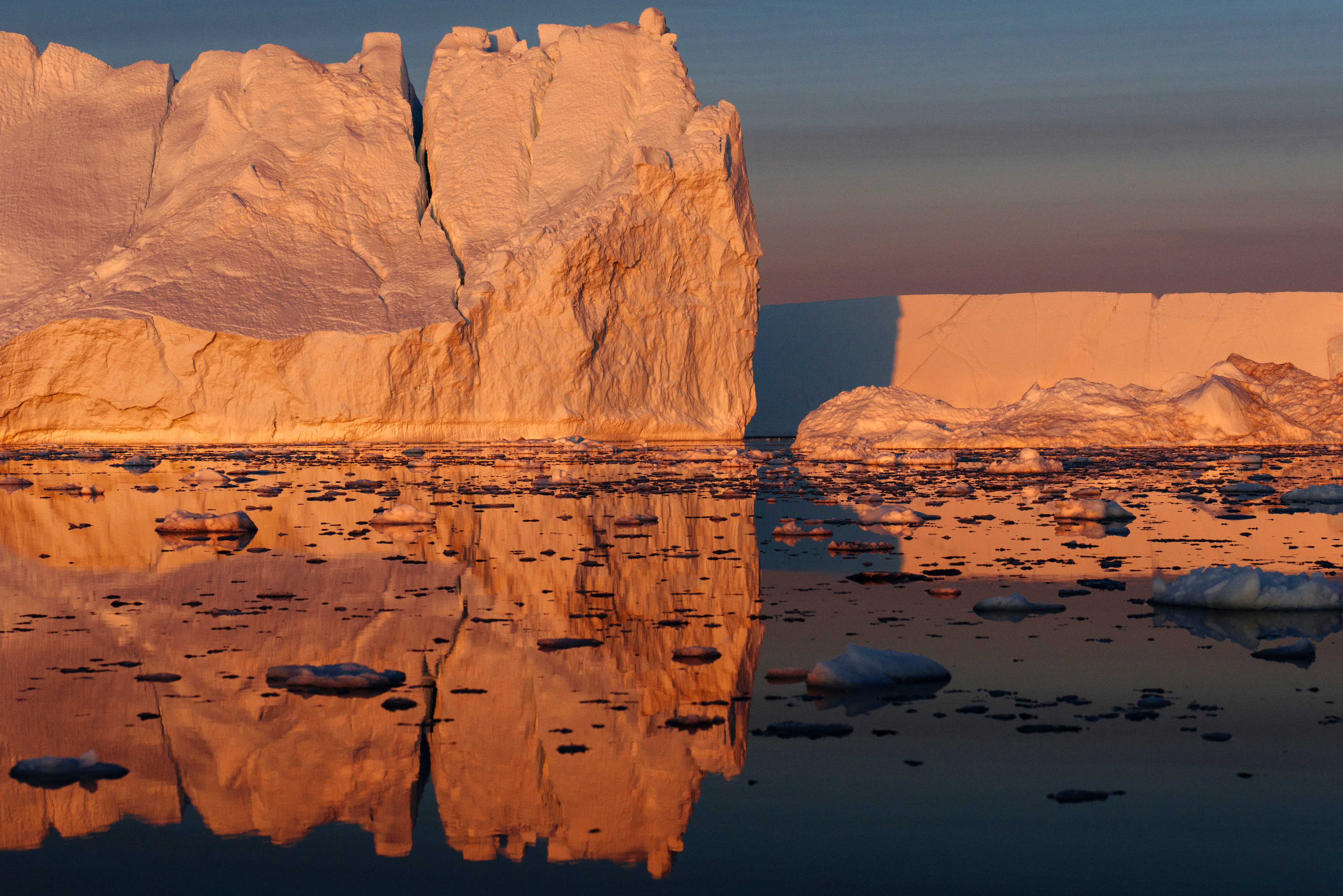 Um imenso bloco de gelo flutuante reflete tons alaranjados e rosados da luz do pôr do sol em águas calmas do Ártico. Fragmentos menores de gelo estão espalhados pela superfície, criando um espelho natural que duplica as cores quentes no frio ambiente polar. A cena transmite a grandiosidade e a fragilidade das geleiras diante das mudanças climáticas.