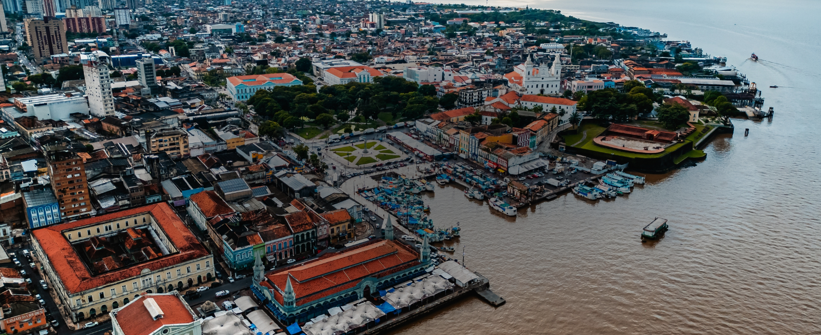 Vista aérea de Belém, cidade histórica às margens da baía do Guajará e que irá receber a COP30 em 2025. A imagem mostra uma área urbana densa, com prédios de arquitetura colonial e telhados vermelhos predominantes. Em destaque, o Mercado Ver-o-Peso, um marco cultural e turístico, com sua estrutura coberta e movimentação de barcos e pequenas embarcações no cais. Próximo ao mercado, áreas verdes bem cuidadas, praças e um forte histórico reforçam o aspecto cultural do local. O rio, imponente, se estende até o horizonte, contrastando com o céu nublado que cobre a cena.