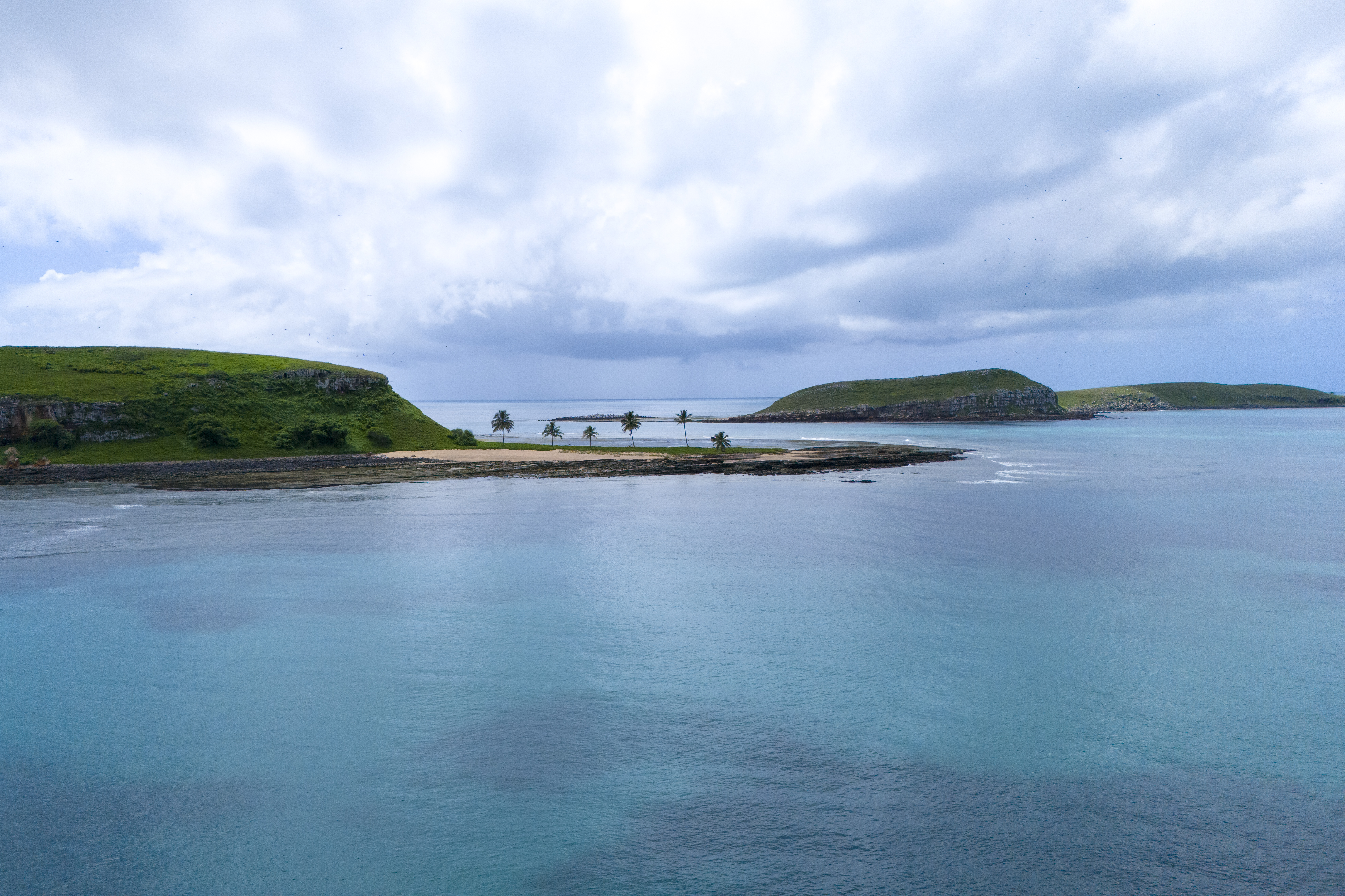 Paisagem costeira com mar calmo, ilhotas rochosas cobertas por vegetação e uma estreita faixa de terra com coqueiros, evidenciando a beleza natural e o equilíbrio ambiental do Parque Nacional Marinho dos Abrolhos do extremo sul da Bahia.
