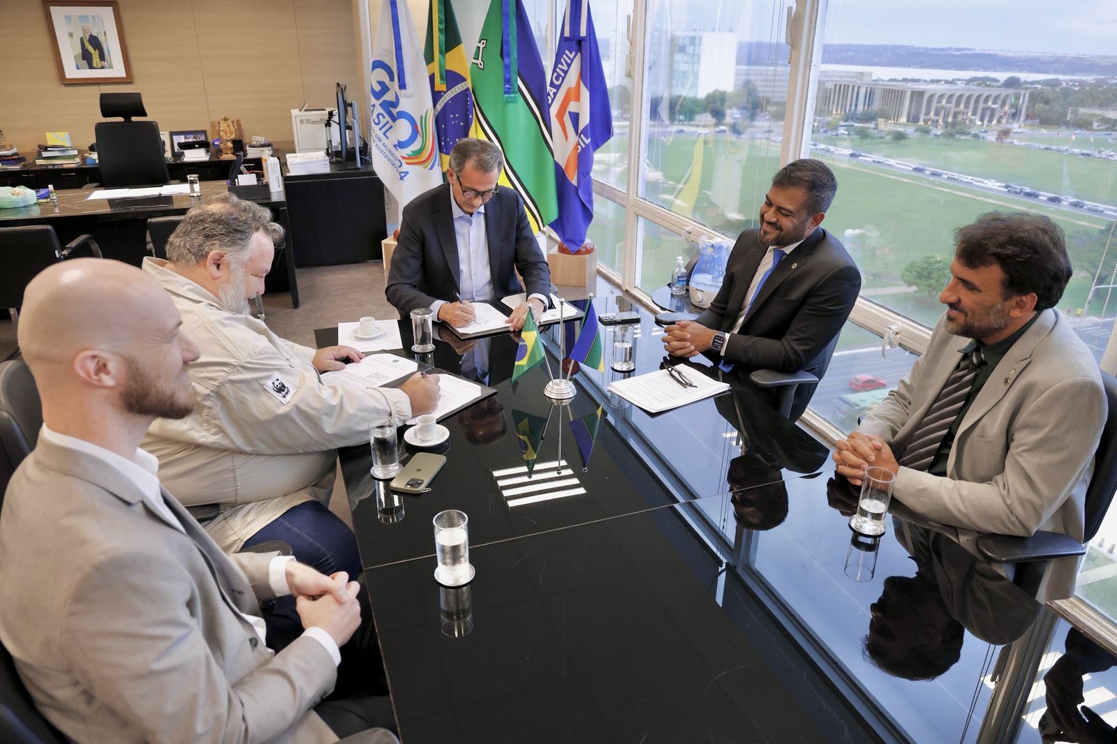 Homens sentados em uma sala de reunião, sendo que dois deles assinam um documento de parceria entre as instituições