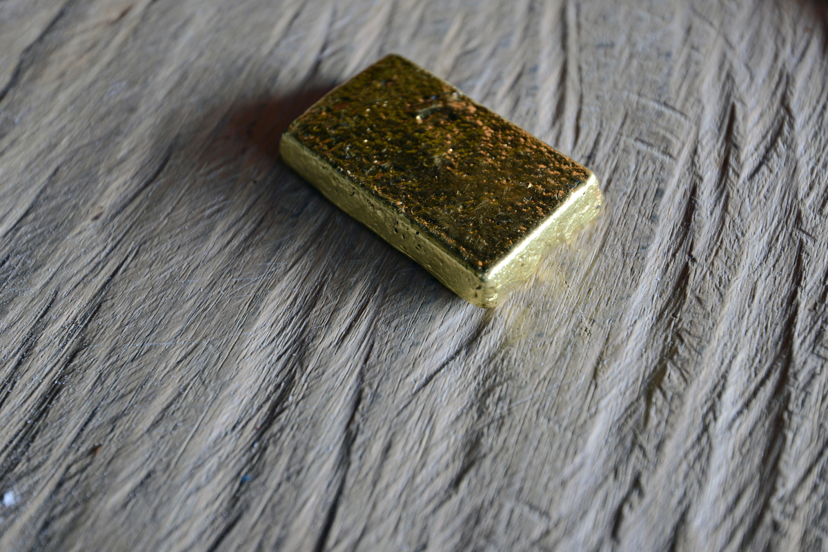 A imagem mostra uma pequena barra retangular de ouro posicionada sobre um tecido de cor cinza-claro, com textura visível e dobras suaves. A superfície do ouro apresenta irregularidades e marcas, indicando um acabamento bruto, não polido. A iluminação lateral cria reflexos dourados e sombras suaves ao redor da peça, destacando o contraste entre o brilho metálico do ouro e o fundo opaco do tecido. Não há outros objetos ou elementos visíveis na cena.